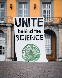 A large banner is displayed in front of a building with columns and arched windows. The banner reads 'UNITE behind the SCIENCE' in bold letters and features a circular green emblem at the bottom with the text 'FRIDAYS FOR FUTURE Bonn' and an image of the Earth.