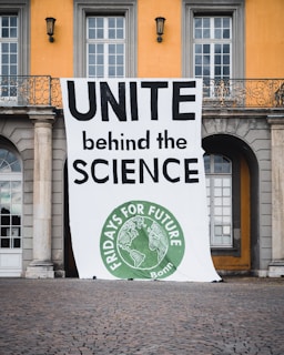 A large banner is displayed in front of a building with columns and arched windows. The banner reads 'UNITE behind the SCIENCE' in bold letters and features a circular green emblem at the bottom with the text 'FRIDAYS FOR FUTURE Bonn' and an image of the Earth.