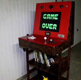 An arcade gaming cabinet with a wooden exterior and a red control panel is prominently displayed. The screen shows the text 'GAME OVER' in green letters against a dark background. The control panel features two joysticks and several white and black buttons. Below the screen, shelves are filled with books, and a small Pikachu figure can be seen at the bottom. The background includes a white-paneled wall.