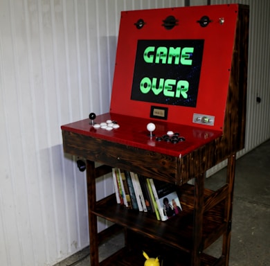 An arcade gaming cabinet with a wooden exterior and a red control panel is prominently displayed. The screen shows the text 'GAME OVER' in green letters against a dark background. The control panel features two joysticks and several white and black buttons. Below the screen, shelves are filled with books, and a small Pikachu figure can be seen at the bottom. The background includes a white-paneled wall.