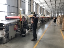A large industrial facility with rows of machinery along the length of the room. Two people are taking photos, one closer to the machines and another in the background. The environment is well-lit with overhead lights, and there are boxes stacked to the side.
