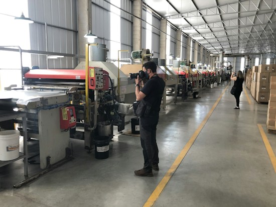 A large industrial facility with rows of machinery along the length of the room. Two people are taking photos, one closer to the machines and another in the background. The environment is well-lit with overhead lights, and there are boxes stacked to the side.