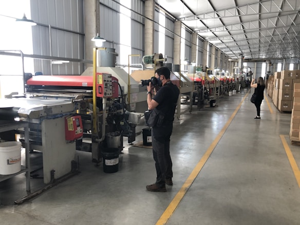 A large industrial facility with rows of machinery along the length of the room. Two people are taking photos, one closer to the machines and another in the background. The environment is well-lit with overhead lights, and there are boxes stacked to the side.