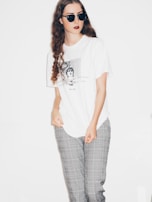 A person with long, curly brown hair wearing black sunglasses, a white graphic t-shirt, and gray plaid pants. The background is plain white, highlighting the clothing and accessories.