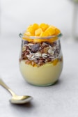 A jar filled with a layered dessert consisting of creamy pudding at the bottom, topped with granola and chunks of yellow fruit. Beside the jar, a spoon rests on a light-colored surface.
