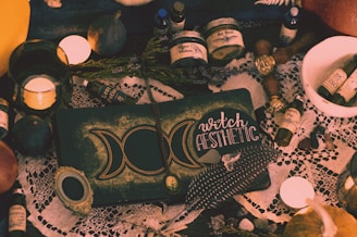 A collection of mystical items arranged on a table, including candles, bottles labeled with potions, a book with the words 'witch aesthetic', feathers, dried herbs, and various small stones. The setting has a vintage and magical feel, with a lace cloth adding an ornate touch to the scene.