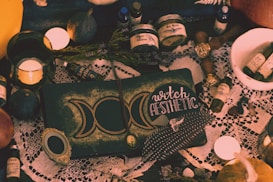 A collection of mystical items arranged on a table, including candles, bottles labeled with potions, a book with the words 'witch aesthetic', feathers, dried herbs, and various small stones. The setting has a vintage and magical feel, with a lace cloth adding an ornate touch to the scene.