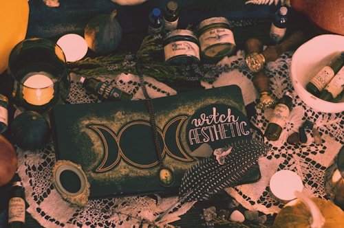 A collection of mystical items arranged on a table, including candles, bottles labeled with potions, a book with the words 'witch aesthetic', feathers, dried herbs, and various small stones. The setting has a vintage and magical feel, with a lace cloth adding an ornate touch to the scene.
