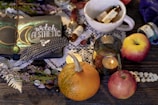 A cozy arrangement featuring a notebook with 'witch aesthetic' written on it, adorned with a moon and feathers. Surrounding the notebook are various items including a small pumpkin, apples, a lit candle in a glass holder, and small bottles with handwritten labels. These items rest on a wooden table with lace and leafy elements, creating an enchanting and mystical ambiance.