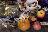 A cozy arrangement featuring a notebook with 'witch aesthetic' written on it, adorned with a moon and feathers. Surrounding the notebook are various items including a small pumpkin, apples, a lit candle in a glass holder, and small bottles with handwritten labels. These items rest on a wooden table with lace and leafy elements, creating an enchanting and mystical ambiance.