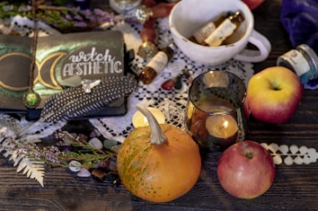 A cozy arrangement featuring a notebook with 'witch aesthetic' written on it, adorned with a moon and feathers. Surrounding the notebook are various items including a small pumpkin, apples, a lit candle in a glass holder, and small bottles with handwritten labels. These items rest on a wooden table with lace and leafy elements, creating an enchanting and mystical ambiance.