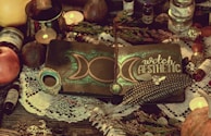 A mystical setup featuring a notebook with a crescent moon design and the words 'witch aesthetic' on the cover. The notebook is adorned with a decorative feather, a crystal, and a pendant. Surrounding objects include candles, an apple, various potion bottles, and a lace cloth, creating an enchanting atmosphere.