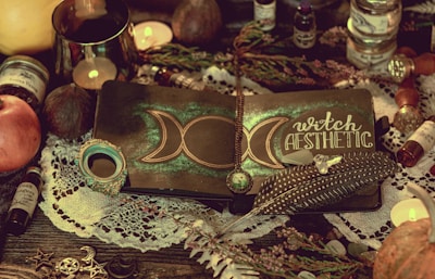 A mystical setup featuring a notebook with a crescent moon design and the words 'witch aesthetic' on the cover. The notebook is adorned with a decorative feather, a crystal, and a pendant. Surrounding objects include candles, an apple, various potion bottles, and a lace cloth, creating an enchanting atmosphere.