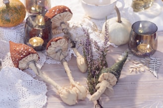 A cozy autumn-themed arrangement featuring decorative mushrooms, a small pumpkin, heather sprigs, and tea light candles in glass holders. The items are placed on a wooden surface adorned with lace doilies and dried leaves, creating a rustic and warm atmosphere.