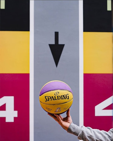 A hand holds a yellow and purple Spalding basketball in front of a colorful sports court or mural, featuring an arrow pointing downwards and the number 4 on both sides. The background is divided into bold sections of black, yellow, red, and gray.