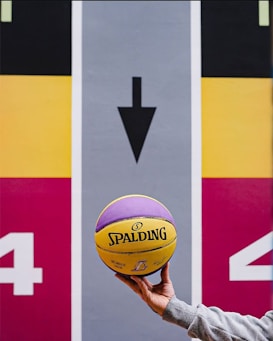 A hand holds a yellow and purple Spalding basketball in front of a colorful sports court or mural, featuring an arrow pointing downwards and the number 4 on both sides. The background is divided into bold sections of black, yellow, red, and gray.
