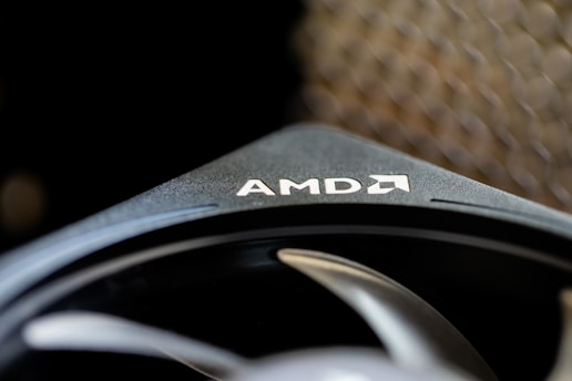 A close-up of a component with the AMD logo prominently visible on a sleek black surface. The image captures the texture and design of the component, highlighting its modern and industrial aesthetic. In the background, a blurred pattern adds depth to the composition.
