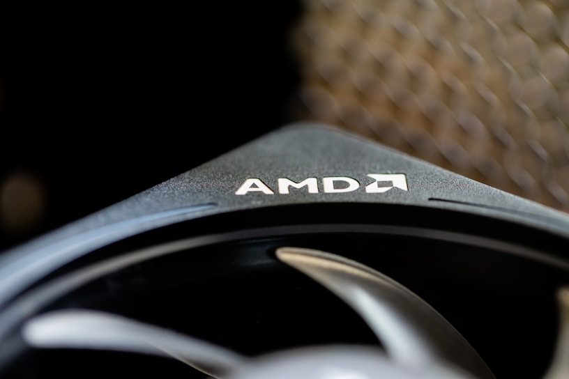 A close-up of a component with the AMD logo prominently visible on a sleek black surface. The image captures the texture and design of the component, highlighting its modern and industrial aesthetic. In the background, a blurred pattern adds depth to the composition.