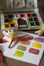 A watercolor palette sits open on a table, displaying a variety of bright pigments. In the foreground, a piece of watercolor paper holds several painted swatches in colors like green, yellow, and orange. A plumeria flower, with soft pink and white petals, rests on the paper alongside a couple of fallen leaves.
