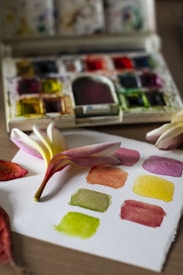 A watercolor palette sits open on a table, displaying a variety of bright pigments. In the foreground, a piece of watercolor paper holds several painted swatches in colors like green, yellow, and orange. A plumeria flower, with soft pink and white petals, rests on the paper alongside a couple of fallen leaves.