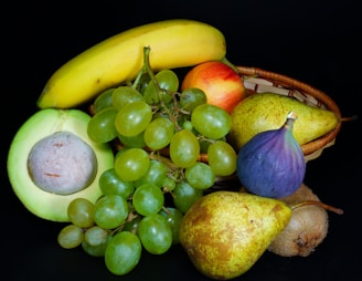 A vibrant assortment of fresh fruits displayed against a dark background. The arrangement includes a banana, grapes, an avocado, pear, kiwi, apple, and fig, some of which are nestled in a wicker basket.