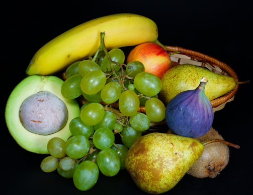 A vibrant assortment of fresh fruits displayed against a dark background. The arrangement includes a banana, grapes, an avocado, pear, kiwi, apple, and fig, some of which are nestled in a wicker basket.