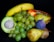 A vibrant assortment of fresh fruits displayed against a dark background. The arrangement includes a banana, grapes, an avocado, pear, kiwi, apple, and fig, some of which are nestled in a wicker basket.