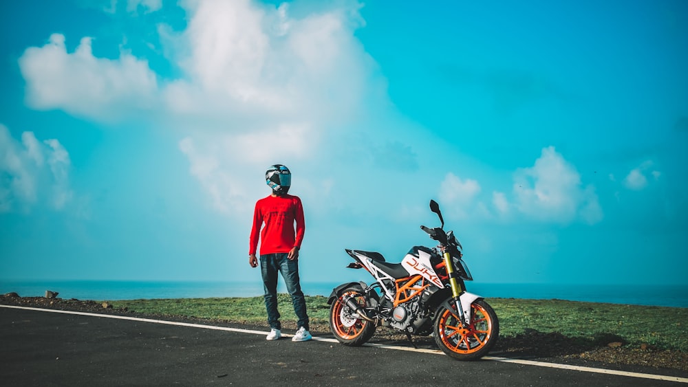 man in red jacket and blue denim jeans standing beside black and white motorcycle during daytime