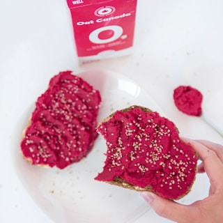 Two slices of bread are spread with a vibrant red beet hummus and sprinkled with sesame seeds. A hand is holding one slice, showing a bite taken out of it. A carton of oat milk is in the background, partially visible. The scene is set on a white plate and table, with a spoonful of hummus nearby.
