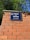 A blue street sign with white text that reads 'Avinguda de Madrid, Capital d'Espanya' is mounted on a brick wall. The wall is made of red bricks with visible mortar lines. Green foliage from trees is visible above the wall, against a clear blue sky.