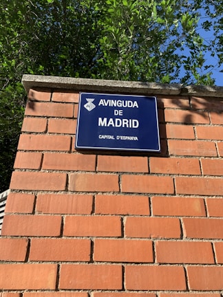 A blue street sign with white text that reads 'Avinguda de Madrid, Capital d'Espanya' is mounted on a brick wall. The wall is made of red bricks with visible mortar lines. Green foliage from trees is visible above the wall, against a clear blue sky.