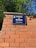 A blue street sign with white text that reads 'Avinguda de Madrid, Capital d'Espanya' is mounted on a brick wall. The wall is made of red bricks with visible mortar lines. Green foliage from trees is visible above the wall, against a clear blue sky.