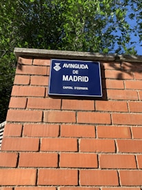 A blue street sign with white text that reads 'Avinguda de Madrid, Capital d'Espanya' is mounted on a brick wall. The wall is made of red bricks with visible mortar lines. Green foliage from trees is visible above the wall, against a clear blue sky.