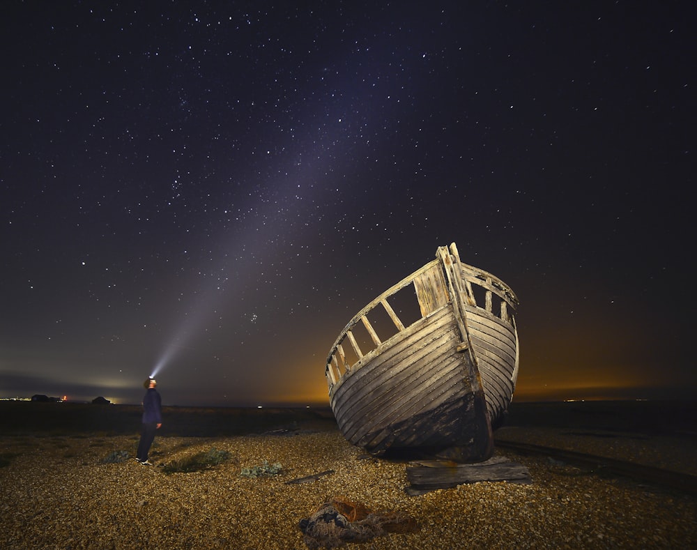 Mann steht neben braunem Holzboot unter sternenklarer Nacht