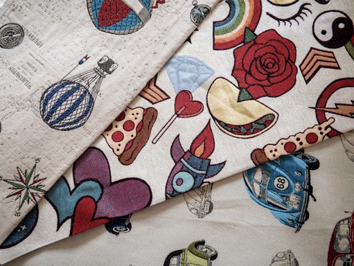 A collection of fabric swatches featuring various colorful designs. The patterns include a stylized hot air balloon, a rose, a rainbow, yin-yang symbol, pizza slice, a heart with flames, vintage cars, and other assorted graphics. The designs are vibrant and eclectic, with a mix of traditional and pop culture elements.