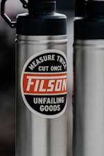 A stainless steel water bottle features a prominent label with the words 'Measure Twice, Cut Once, FILSON, Unfailing Goods' in a circle. The background is blurred, highlighting the bottle's design and text.