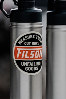 A stainless steel water bottle features a prominent label with the words 'Measure Twice, Cut Once, FILSON, Unfailing Goods' in a circle. The background is blurred, highlighting the bottle's design and text.