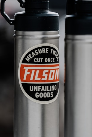 A stainless steel water bottle features a prominent label with the words 'Measure Twice, Cut Once, FILSON, Unfailing Goods' in a circle. The background is blurred, highlighting the bottle's design and text.