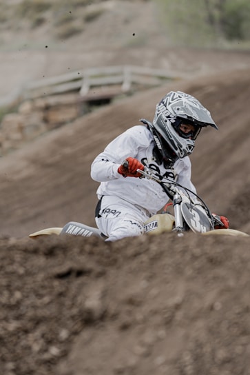 A motorcyclist in full racing gear is navigating a dirt track on an off-road motorcycle. The rider is wearing a helmet and gloves, and there is dirt being kicked up from the track as they maneuver the bike. The background shows a blurred view of a dirt track.