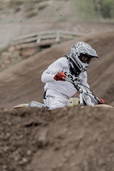 A motorcyclist in full racing gear is navigating a dirt track on an off-road motorcycle. The rider is wearing a helmet and gloves, and there is dirt being kicked up from the track as they maneuver the bike. The background shows a blurred view of a dirt track.