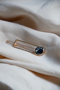 A round black and white marbled brooch with a gold rim is placed on a soft, light beige fabric surface. The fabric appears smooth and slightly wrinkled, creating a gentle, flowing texture around the brooch.