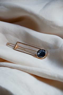 A round black and white marbled brooch with a gold rim is placed on a soft, light beige fabric surface. The fabric appears smooth and slightly wrinkled, creating a gentle, flowing texture around the brooch.