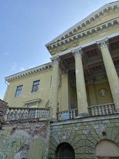 A classical style building with tall columns, intricate moldings, and a decorative frieze. The facade shows signs of decay with peeling paint and exposed brickwork. The architecture includes balustrades and a relief portrait set into the wall.
