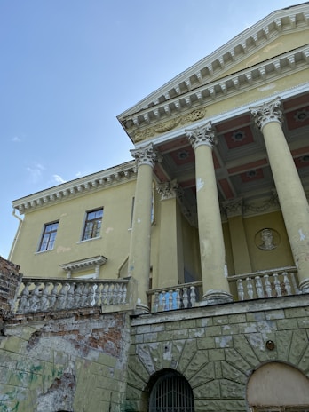 A classical style building with tall columns, intricate moldings, and a decorative frieze. The facade shows signs of decay with peeling paint and exposed brickwork. The architecture includes balustrades and a relief portrait set into the wall.
