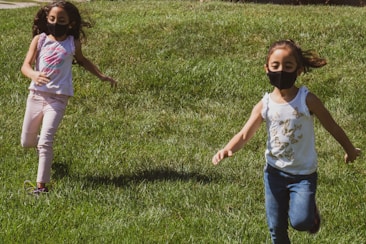 Two young children wearing face masks are running joyfully on a green lawn. The sunlight casts shadows on the grass, and both children are dressed casually in summery outfits.