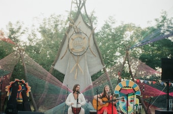 Two musicians are performing on an outdoor stage with colorful decorations and artistic backdrops. The setup includes a large triangular structure with tribal motifs and yarn art. A man plays a drum, while a woman plays an acoustic guitar. The natural surroundings include trees and open sky.
