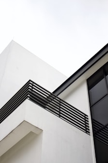 A modern architectural structure with clean lines featuring a white building facade and a balcony with horizontal metal railings. The design is minimalist with large glass windows and a flat roof.