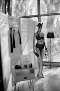 A mannequin dressed in dark lingerie and a mask stands next to a lamp. Nearby, a small table holds various items, including a sign with text. The setting includes paneled dividers and large windows filtering light.
