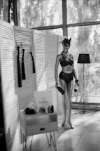 A mannequin dressed in dark lingerie and a mask stands next to a lamp. Nearby, a small table holds various items, including a sign with text. The setting includes paneled dividers and large windows filtering light.