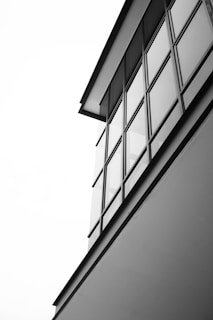 A minimalist black and white photograph of a modern building with large glass windows and clean lines. The structure is captured from a low angle, emphasizing its geometric shapes and sleek design against a stark white sky.